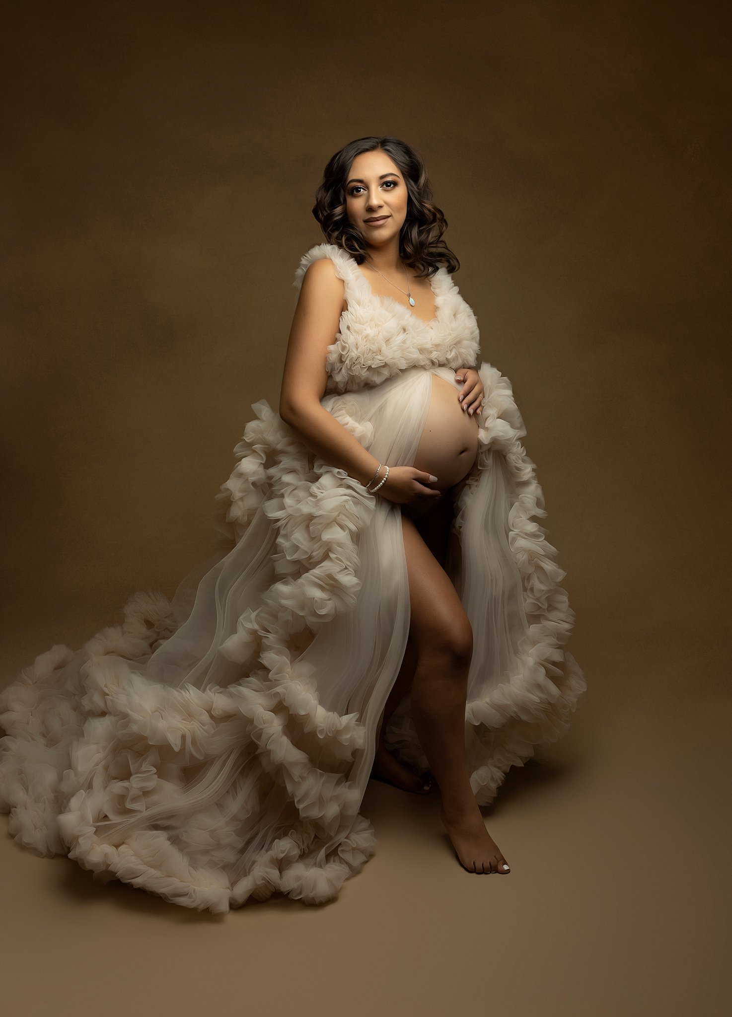 Mother to be stands in a studio wearing a white maternity gown while holding her bump