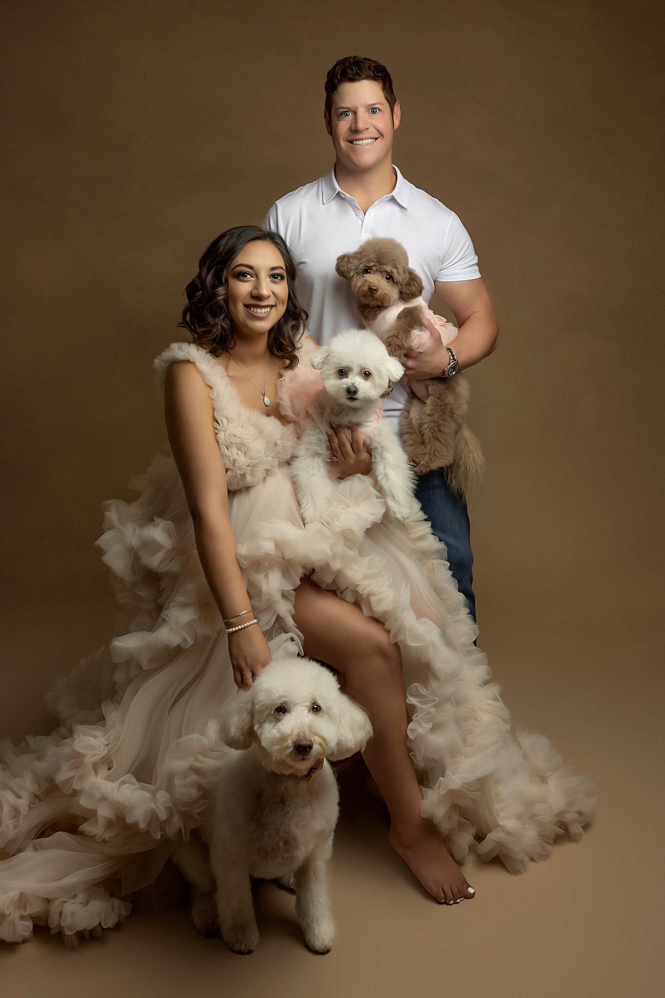 Mother to be in a beige maternity gown sits on a stool in a studio with her partner and three fluffy dogs MUSC birthing center