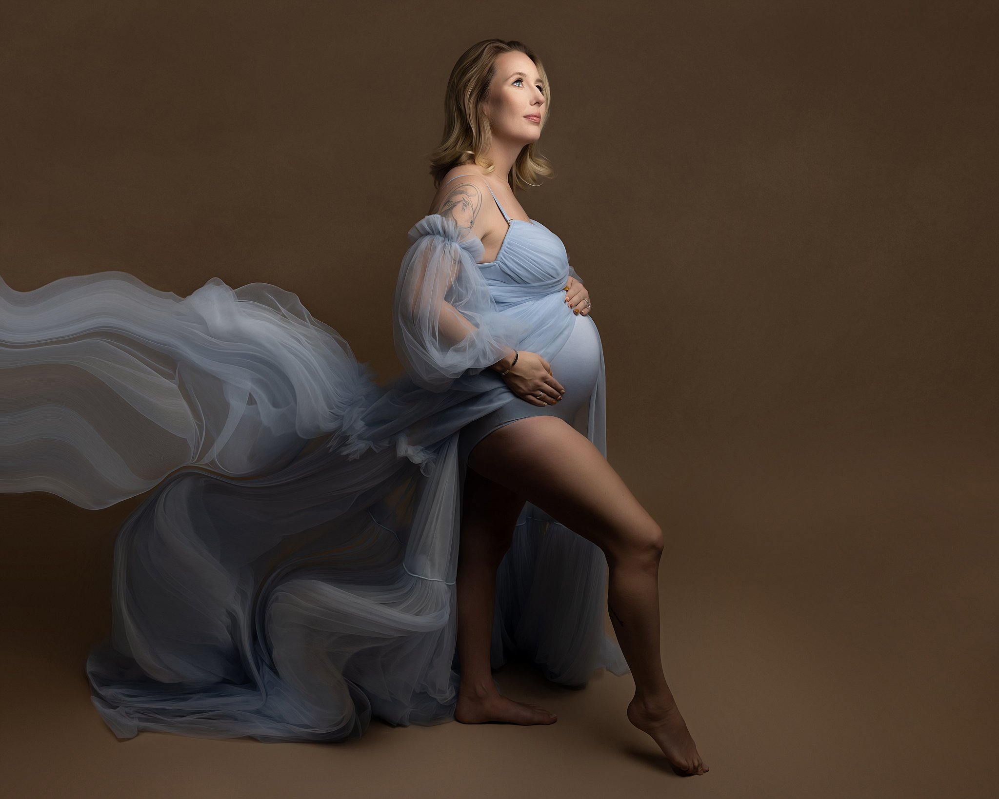 Pregnant woman stands in a studio in a flowing blue maternity gown