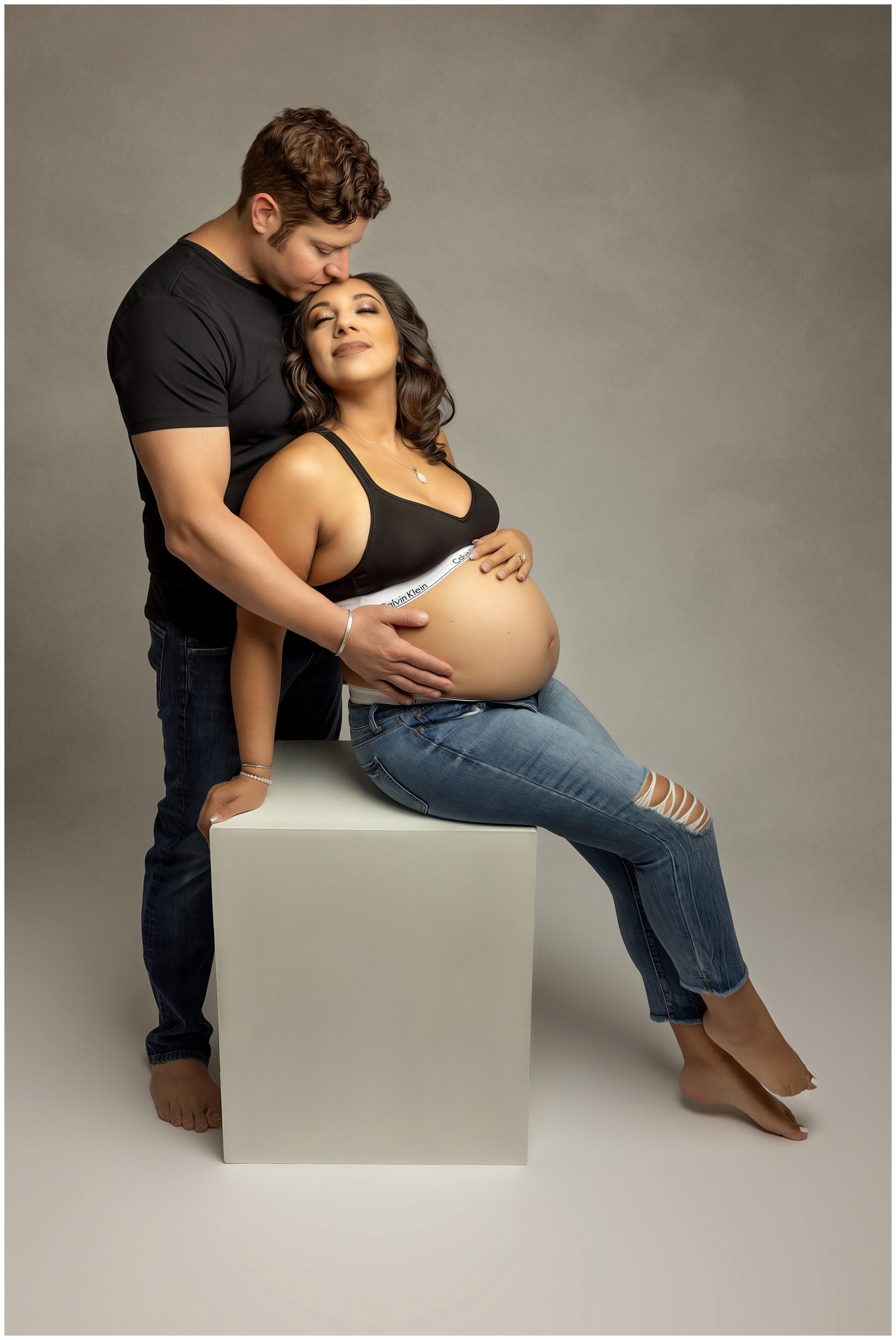 Soon to be dad kisses the forhead and holds the bump of mother to be while sitting on a box in a studio midwives charleston sc