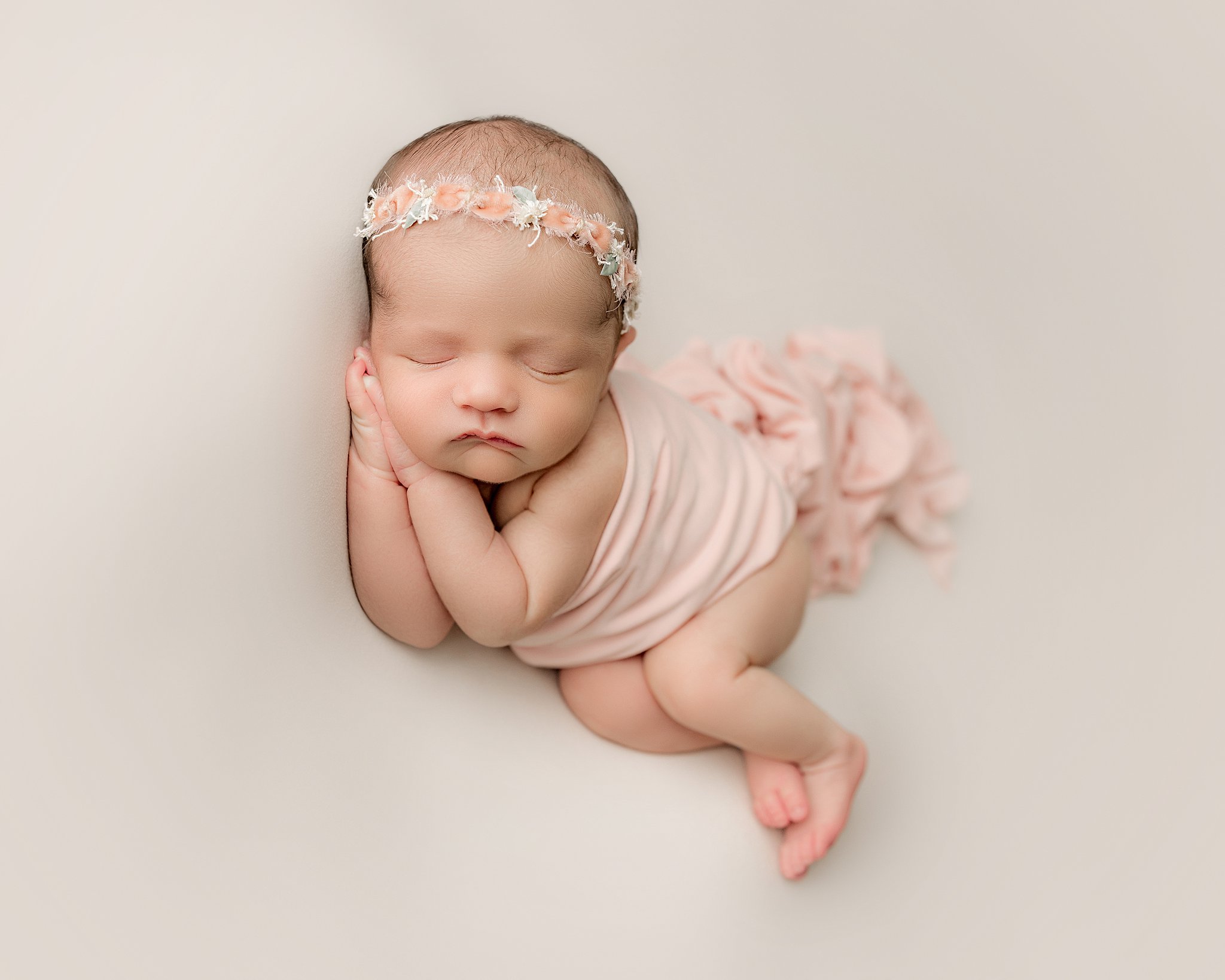 A newborn baby sleeps while resting her head on her hands while wrapped in a pink blanket and a floral headband baby bloomers charleston