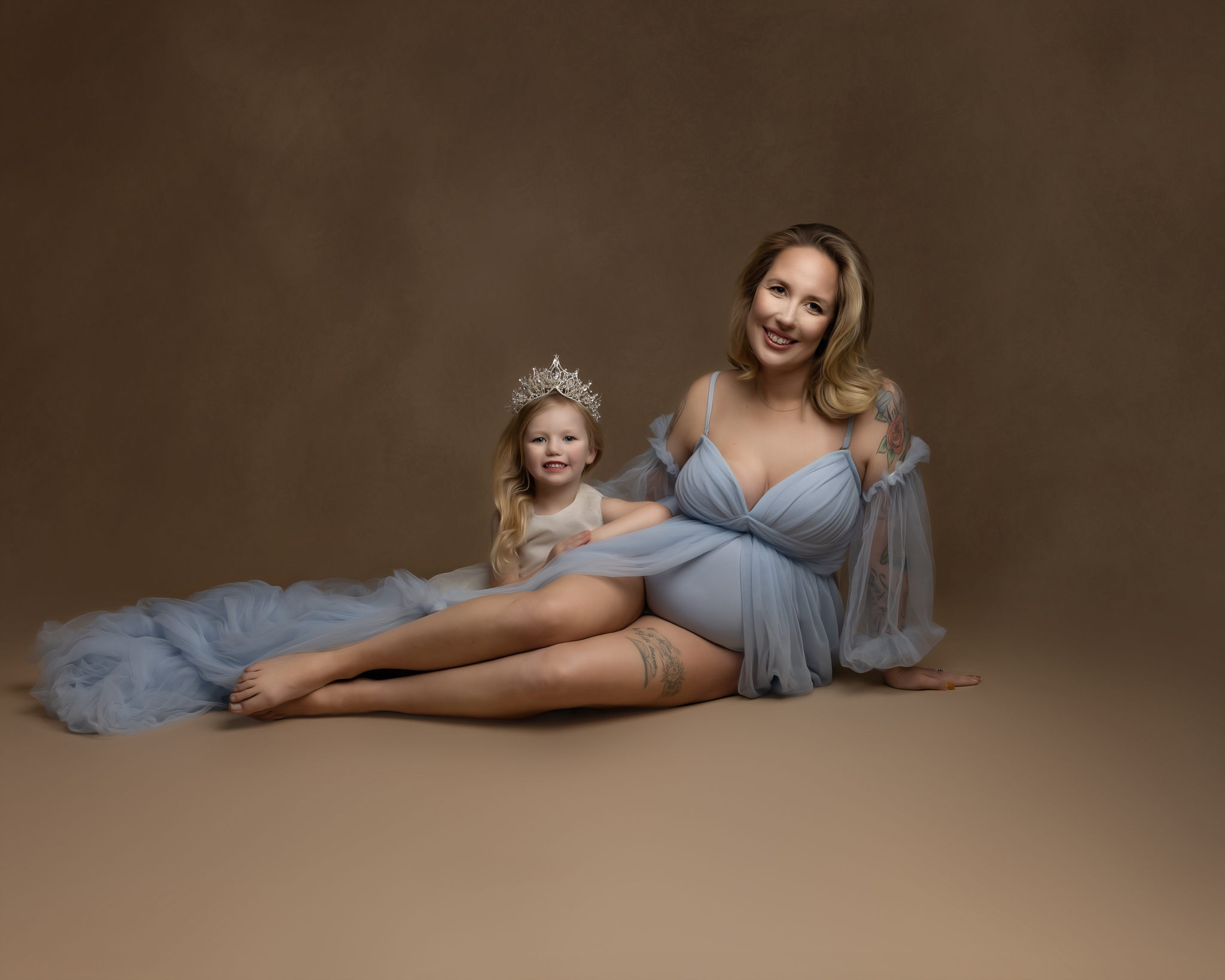 A mother to be in a blue maternity gown sits on the floor of a studio with her toddler daughter wearing a princess tiara at her side charleston obgyn