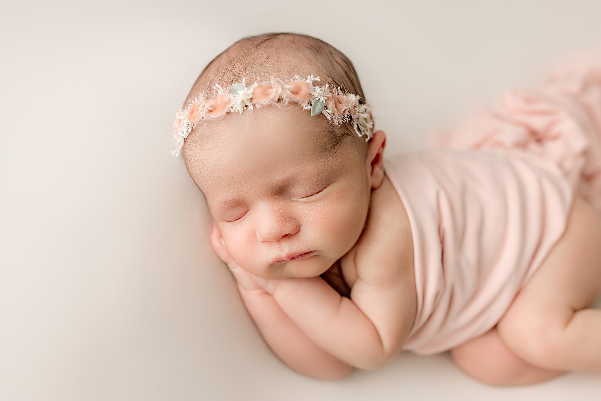 A newborn baby wrapped in a pink blanket sleeps on its hands wearing a floral headband lowcountry baby boutique