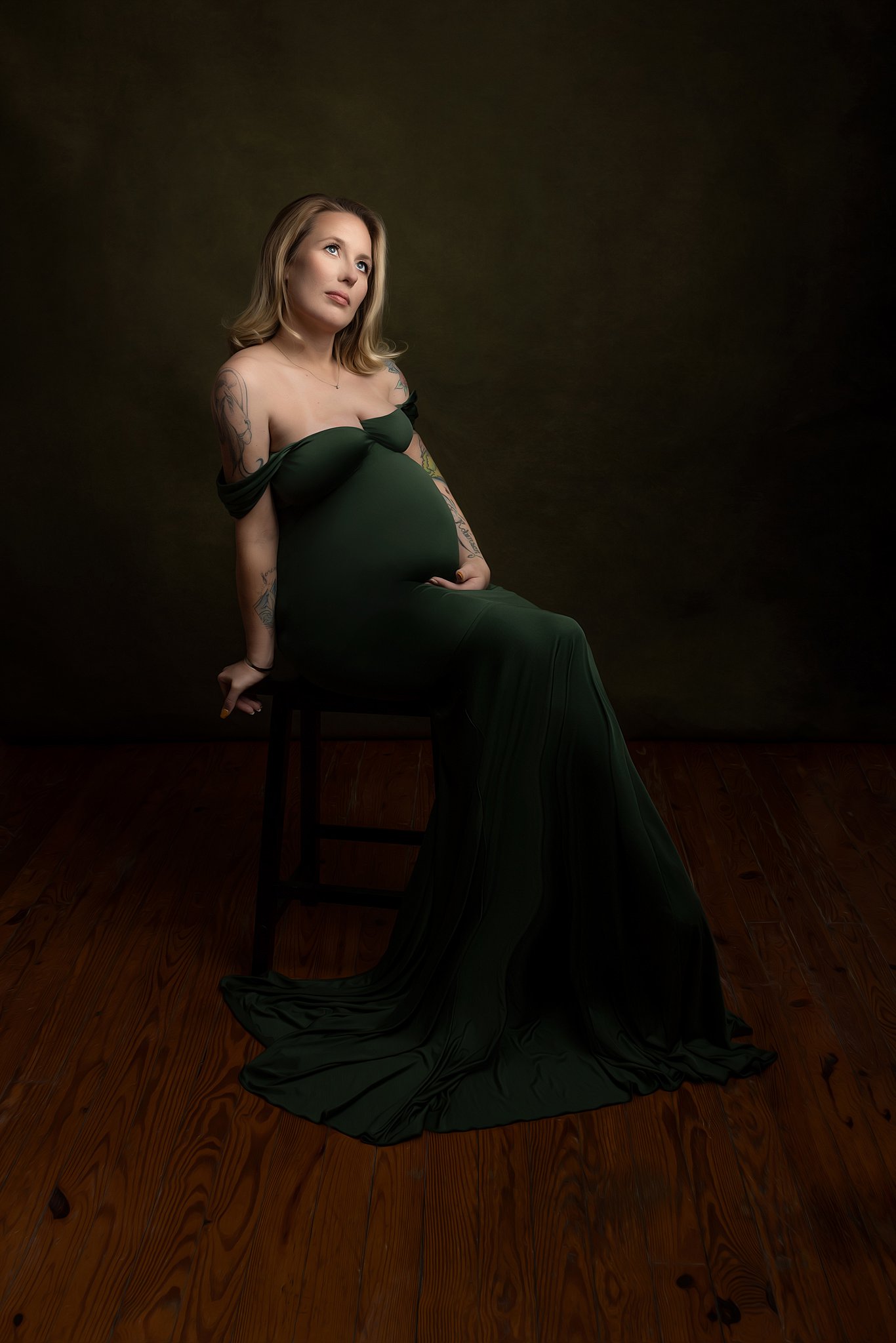 A pregnant woman sits on a dark wooden chair in a studio while wearing a green maternity gown off her shoulders