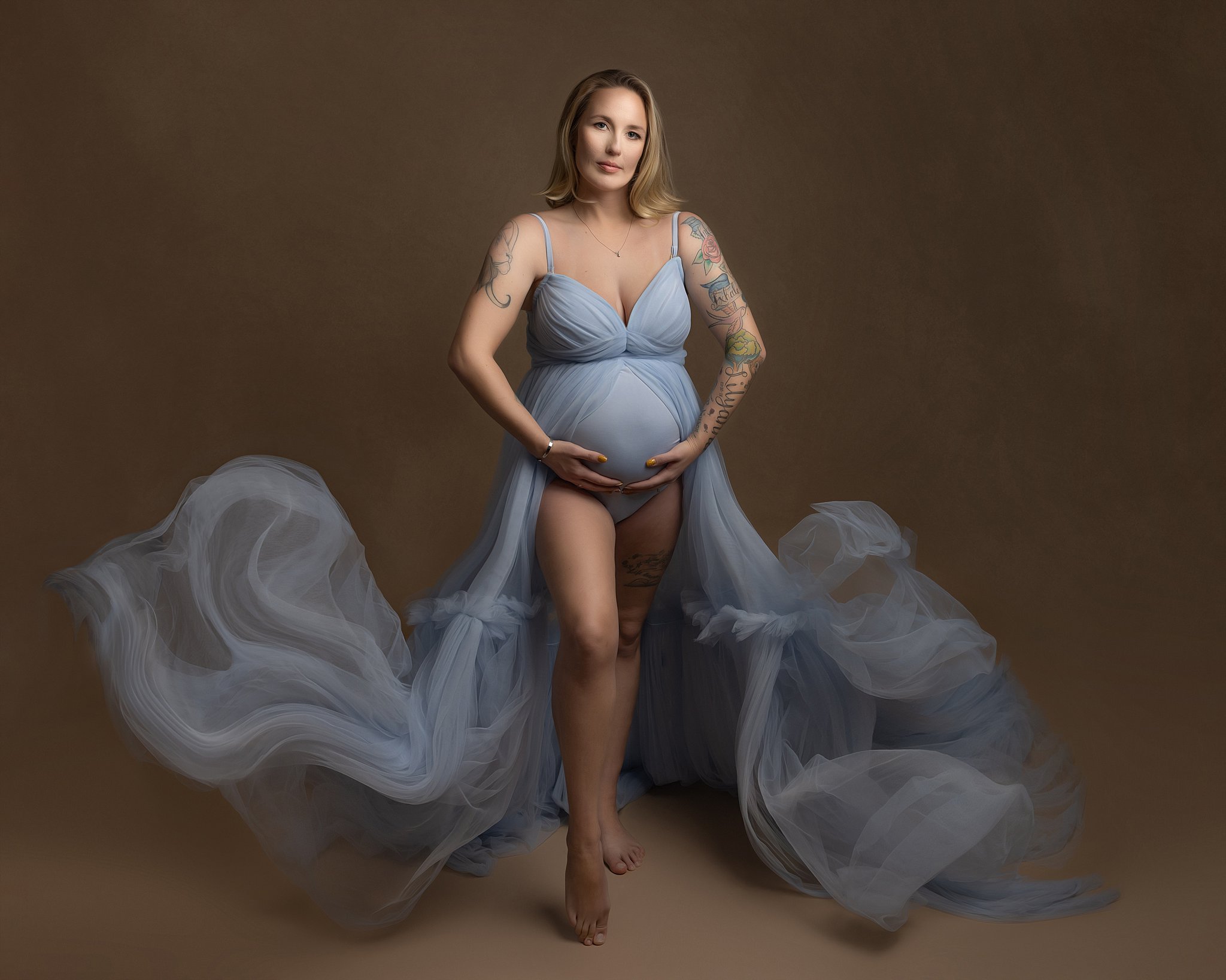 A mother to be in a blue maternity gown stands in a studio holding her bump with both hands while her train flows around her prenatal massage charleston sc
