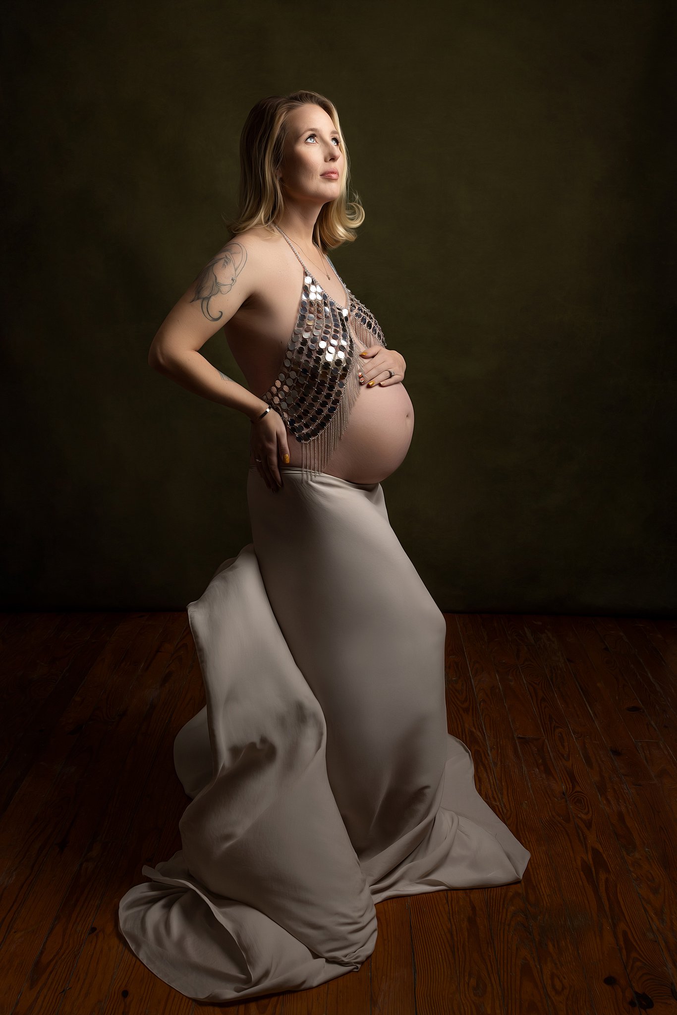 A mom to be in a shiny top and flowing dress stands in a studio with a hand on her bump and one on her back prenatal massage charleston sc