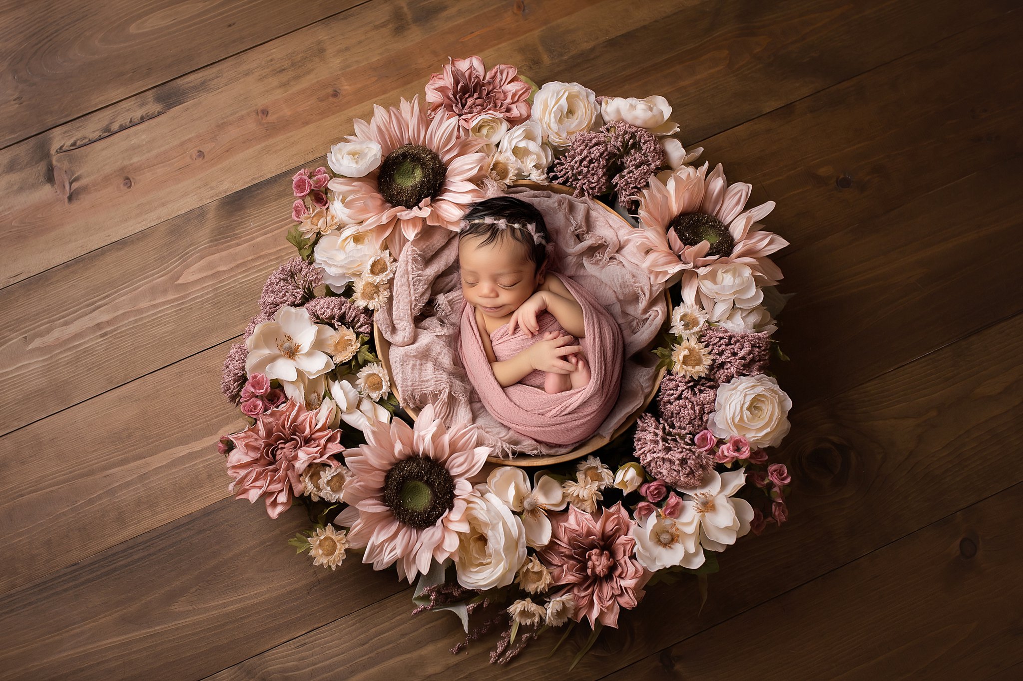 A sleeping newborn baby lays in a wooden bucket in a wreath of flowers